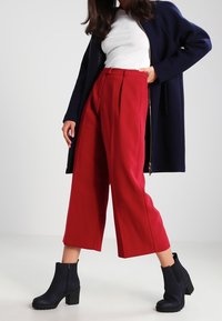 Pantaloni a vita alta rossi, larghi e con pieghe, abbinati a un top in maglia bianco e a un lungo cappotto blu navy. Stivaletti neri con tacco completano l'outfit.