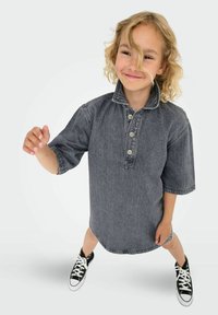 Otrok s kodrastimi blond lasmi, oblečen v prevelik temno modri jeans srajci in črnimi športnimi copati, se smeška in stoji z narazen postavljenimi nogami na bež ozadju.