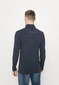 Laivastonsininen pitkähihainen polo-paita, jossa on korkea kaulus. Takana on minimalistinen printti. Pehmeä kangas, jolla on sileä rakenne.