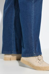 Jambes portant un jean en denim bleu et des chaussures en toile beige avec des semelles en corde tressée, debout sur une surface blanche avec un fond gris clair.