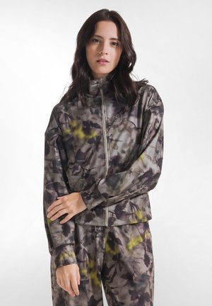 Joven mujer con cabello oscuro lleva una chaqueta y pantalones con cierre, a juego, de patrón floral gris y amarillo, de pie frente a un fondo blanco liso.