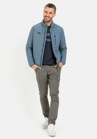 Hellblau gesteppte Jacke mit Stehkragen, Reißverschlusstasche, getragen über einem marineblauen T-Shirt, kombiniert mit grauen Cargo-Hosen und weißen Sneakers.