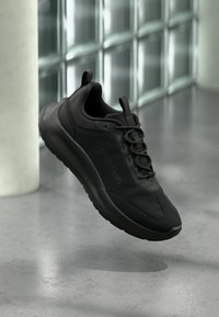 Basket noire Calvin Klein avec lacets, flottant au-dessus d'un sol gris près d'un mur en blocs de verre et d'une colonne blanche en arrière-plan.