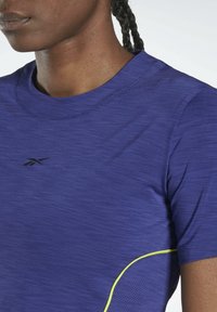 T-shirt de sport violet en tissu léger et respirant. Il présente un col rond, un design sans coutures et de subtils accents jaunes sur les côtés.