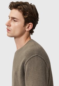 Grijze, gestructureerde trui met een ronde hals, zichtbare stiksels en een klein logo op de borst. De stof lijkt zacht en licht flufachtig.