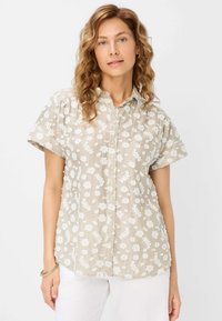 Camicetta a maniche corte in tessuto beige chiaro con ricami floreali bianchi, caratterizzata da una chiusura con bottoni e una vestibilità rilassata.
