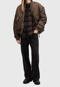 Brun bomberjacka med dragkedja och framfickor, lagd över en rutig skjorta i mörka nyanser. Kombinerad med svarta vida jeans och stövlar.