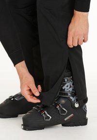 Person som spänner fast en svart skidsko med spännen, justerar svarta byxor med dragkedja och kardborre vid ankeln över skon.