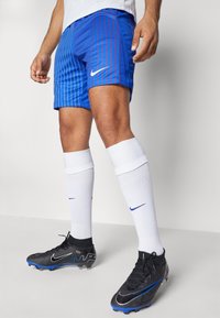 Nike blå sportshorts med röda vertikala ränder, kombinerat med vita knähöga strumpor och svarta fotbollsskor med blå detaljer.