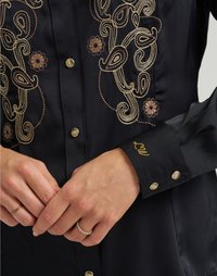 Blusa preta com intrincadas bordaduras em dourado e castanho, botões dourados e um punho com "LW" em fio dourado. Textura semelhante à seda.