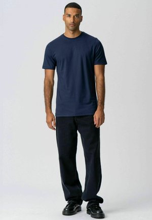 Mann steht und trägt ein marineblaues T-Shirt, schwarze weite Hosen und schwarze Schuhe vor einem einfarbigen hellen Hintergrund.