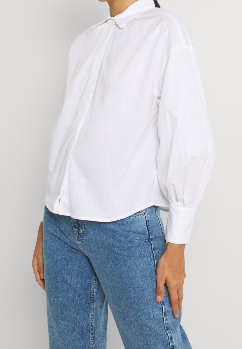 Femme portant une chemise blanche ample à boutons avec des manches longues bouffantes et un jean bleu taille haute, debout devant un fond uni.