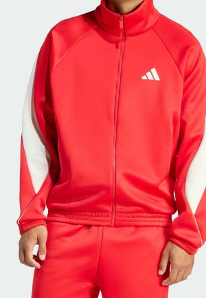 Homme portant une veste de survêtement rouge zippée avec des bandes blanches sur les manches et un pantalon rouge assorti, montrant uniquement le torse et les bras.
