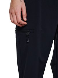 Nærbillede af en hånd hvilende nær en sort bukseben med en lille vandret lynlåslomme.