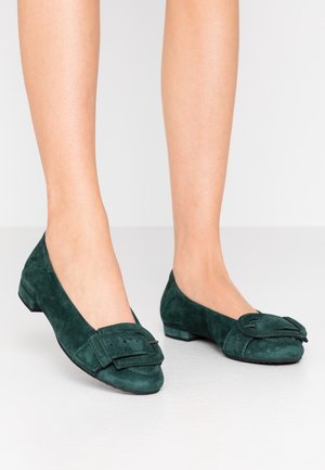 Jambes portant des chaussures plates en daim vert foncé avec une grande boucle décorative à l'avant, debout sur un sol clair devant un fond uni.