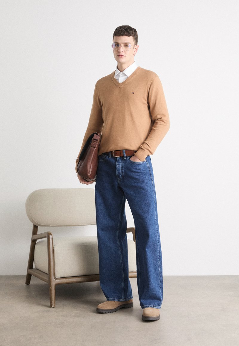 Maglione color cammello con scollo a V, camicia bianca con colletto, jeans blu, cintura marrone, che porta una borsa in pelle marrone, in piedi accanto a una sedia di colore chiaro.