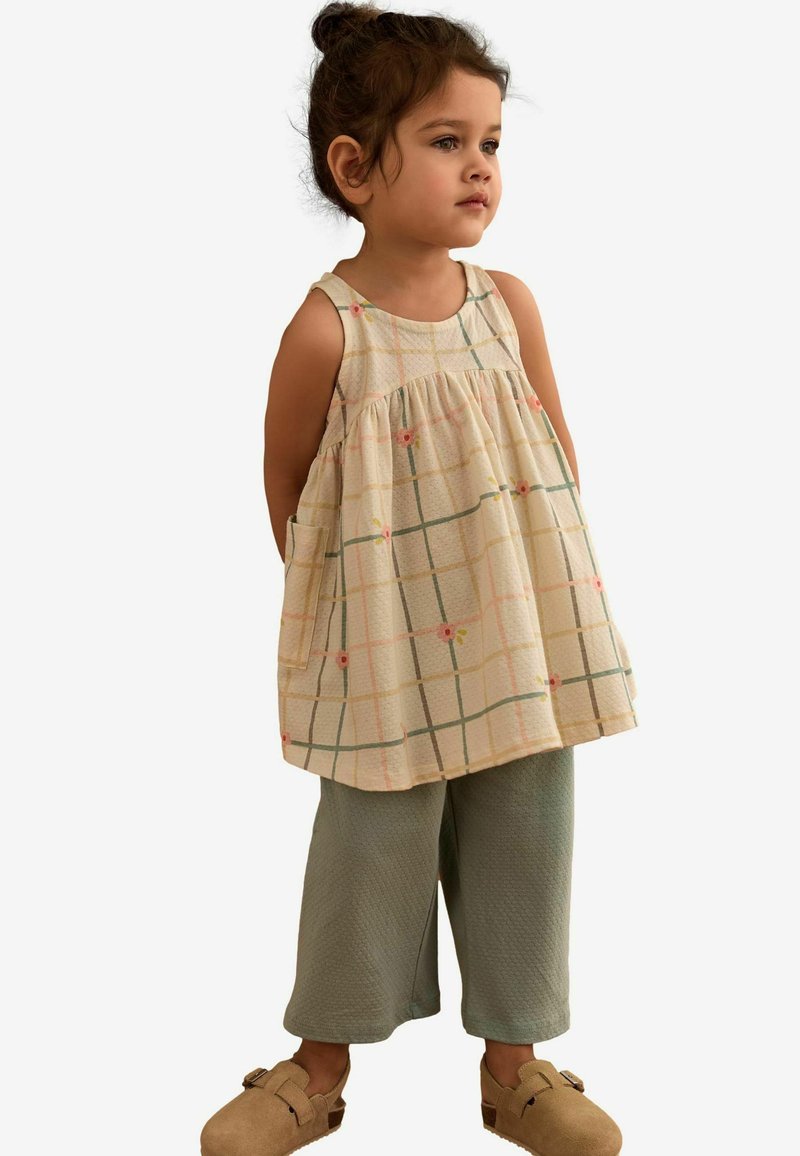 Hellbeige karierte Tanktop mit floralen Akzenten, kombiniert mit grünen strukturierten Culottes und braunen Slippern.