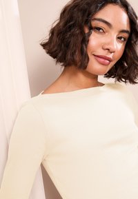 Világos bézs, bordázott, hosszú ujjú felső, széles, csónaknyakkal és puha textúrával, amely kiemeli a visszafogott részleteket a vállrészen.