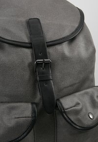 Sac à dos en tissu gris avec sangle boucle en cuir noir et deux poches avant à rabat avec fermetures à boutons.