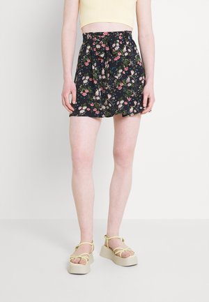 Femme portant une mini-jupe noire à fleurs avec un lien à la taille, un crop top jaune pâle et des sandales compensées avec des brides jaunes.