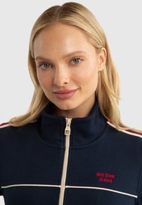 Veste zippée bleu marine avec un col haut, ornée de rayures blanches et rouges sur les épaules et "BIG STAR JEANS" brodé en rouge.