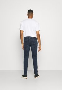 Mies seisoo selin valkoisessa t-paidassa, tummansinisissä farkuissa ja mustissa korkeavartisissa tossuissa yksinkertaista taustaa vasten.