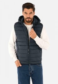 Gilet imbottito blu navy con cappuccio, chiusura zip frontale e due tasche laterali. Presenta un design trapuntato ed è indossato sopra una maglietta bianca a maniche lunghe.