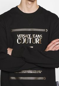 Felpa nera in cotone con testo grafico dorato e design a catena. Presenta un collo a giro e polsini a costine. Il tessuto ha una texture liscia.