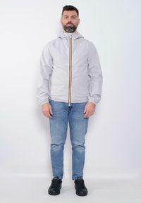 Giacca a zip grigia chiara con cappuccio, caratterizzata da una cerniera con strisce gialle e rosse e polsini elastici; indossata sopra jeans blu e sneakers nere.