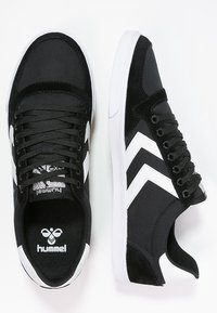 Pari di sneaker Hummel bianche e nere con strisce a chevron, lacci neri e suole bianche, mostrate da vista superiore e laterale.