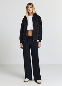 Jeune femme se tenant debout, portant un sweat à capuche noir zippé, un haut court blanc, un pantalon noir à jambes larges et des baskets blanches sur un fond neutre.