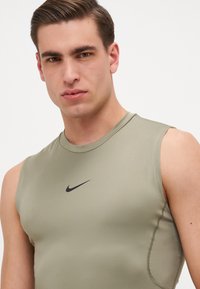 Sportski top bez rukava od maslinasto zelene tkanine, s prilagođenim dizajnom i crnim Nike logotipom na prsima. Glatka tekstura s minimalnim šavovima.