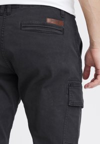 Sorte cargo bukser lavet af tykkere stof, med en baglomme med et lædermærke og en sidelomme med klap. Struktureret overflade.