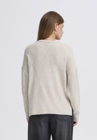 Béžový pletený svetr s diamantovým vzorem, žebrovanou texturou a kulatým výstřihem. Stylizovaný s volnými šedými kalhotami culottes.