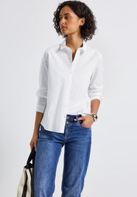 Femme avec des cheveux courts et bouclés, portant une chemise blanche à boutons et un jean bleu, tenant un sac fourre-tout rayé noir et blanc.