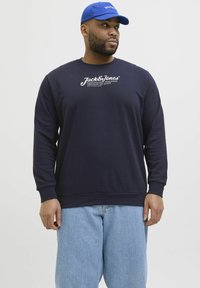 Marineblaue Sweatjacke mit Rundhalsausschnitt und langen Ärmeln, versehen mit weißem, bedrucktem Logotext. Kombiniert mit hellblauen Jeans.