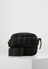 Černá prošívaná crossbody taška se stříbrnými hroty a širokým nastavitelným popruhem na bílém podstavci proti jednoduchému pozadí.