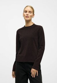 Vrouw met blond haar in een lage paardenstaart, die zilveren oorringen, een donker, langmouwig, textuurtop en zwarte broeken draagt, tegen een effen achtergrond.