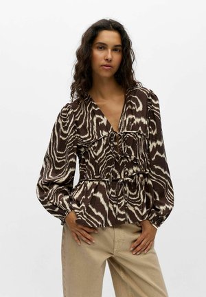 Femme aux longs cheveux bouclés portant un chemisier marron et crème imprimé animalier et un pantalon beige, debout les mains sur les hanches sur fond blanc.