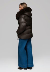 Giacca a vento marrone scuro con un grande colletto in pelliccia sintetica morbida. Il modello indossa pantaloni in denim blu a gamba larga e stivaletti neri, visti da dietro.