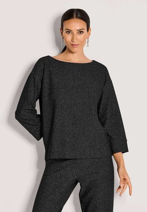 Vrouw met donker haar, draagt hangende oorbellen en een bijpassend zwart textuur langemouwen top en broek, poserend tegen een lichte achtergrond.