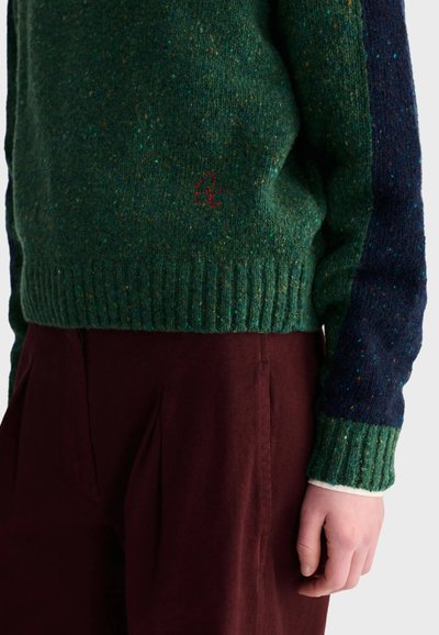 Personne portant un pull en maille vert et marine avec un détail brodé rouge et un pantalon plissé rouge foncé, debout, main droite détendue.
