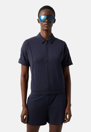 Person iført navyblå kortærmet skjorte med krave og lynlås, matchende shorts og store reflekterende blå solbriller, stående foran en ensfarvet baggrund.