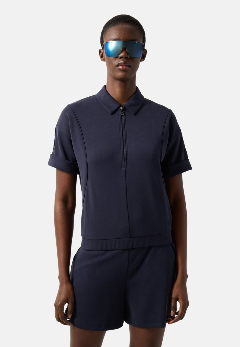 Persoon draagt een marineblauw kortemouwen overhemd met kraag en rits, bijpassende korte broek en grote reflecterende blauwe zonnebril, staand tegen een effen achtergrond.