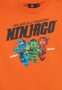 Oranžová košeľa s postavami LEGO Ninjago v modrej, zelenej a červenej farbe, text "Všetci sme draci," "Ninjago" a "Silnejší spolu."