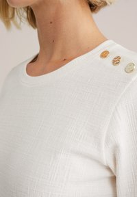 Prim-plan cu o cămașă albă texturată, având trei butoane decorative pe umăr, evidențiind detaliile și designul său fashion.