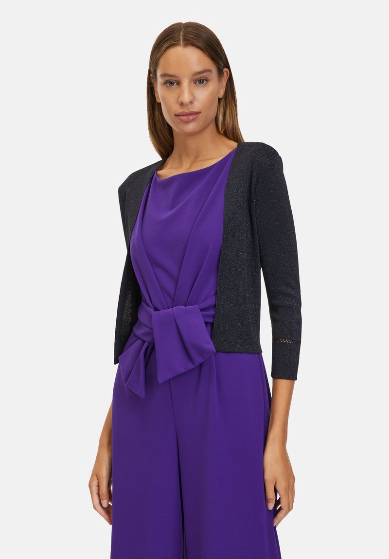 Lila ruha, derékon csomó részlettel, sötét, textúrált kardigánnal párosítva, amely háromnegyedes ujjú és csipke díszítésű.