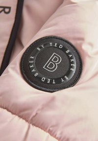 Lyserød polstret stof med et rundt sort mærke med hvid tekst, der står "B" og "by Ted Baker." Tekstureret overflade med syninger.