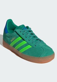 Gröna mockasneakers med tre neongröna ränder, blå detaljer på hälen och en genomskinlig gummisula. Har en klassisk snörningsdesign.