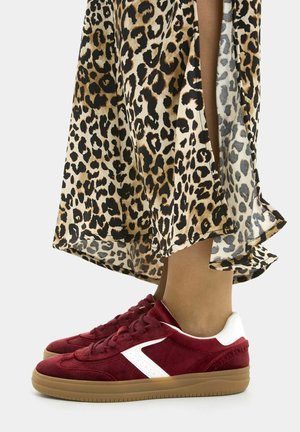 Donkerrode suède sneakers met witte accenten en rubberen zolen, gedragen met een midilengte rok met luipaardprint op een lichte achtergrond.